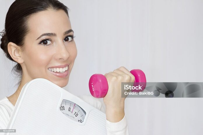 Beautiful young woman holding weight scale and a pink dumbbell in hands.